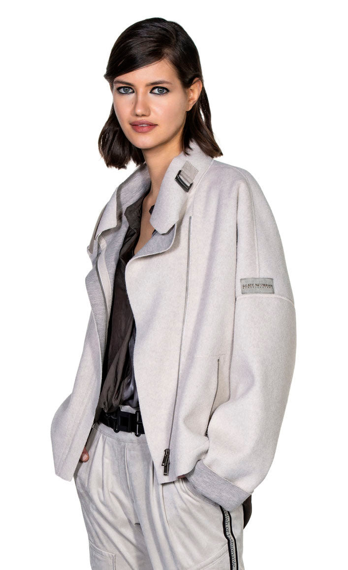 Front top half view of a woman wearing the beate heymann felty jacket in the color sand. This jacket is off-white with a grey lining that shows on the collar and cuffs. The jacket had drop shoulder cuffed long sleeves, an off center zipper front, a stand collar, and a front zip pocket.