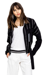 Front, top half view of a woman wearing white pants a white top and the Beate Heymann Stripped Jacket. This jacket is black with white stripes. The sleeves are solid black and the front has two patch, solid black pockets with a single white stripe on them. The jacket has a zip up front and belt near the bottom.