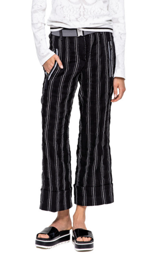 Front, bottom half view of a woman with her hand in the pocket of the Beate Heymann Stripped Culotte Pant. These pants are black with white stripes. They have a straight leg with a cuffed bottom, two front zip pockets, and a black and white belt.