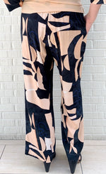 Load image into Gallery viewer, Back bottom half view of a woman wearing the bitte kai rand monstera pant. This pant is nude colored with a black and blue abstract pattern on it. The pants are wide legged.
