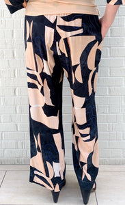 Back bottom half view of a woman wearing the bitte kai rand monstera pant. This pant is nude colored with a black and blue abstract pattern on it. The pants are wide legged.