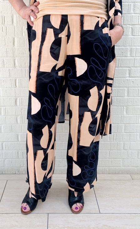 Front bottom half view of a woman wearing the bitte kai rand monstera pant. This pant is nude colored with a black and blue abstract pattern on it. The pants are wide legged.