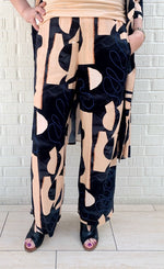 Load image into Gallery viewer, Front bottom half view of a woman wearing the bitte kai rand monstera pant. This pant is nude colored with a black and blue abstract pattern on it. The pants are wide legged.
