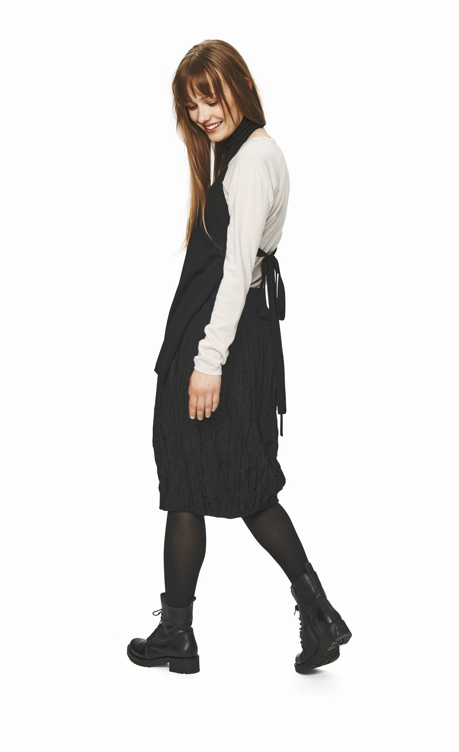 Left side full body view of a woman wearing the bitte kai rand wool front piece in black over a white top and with a black skirt. This front piece has a single front pocket on the bottom right side, no sleeves, and a turtleneck. It is backless with ties that are tied to the back.