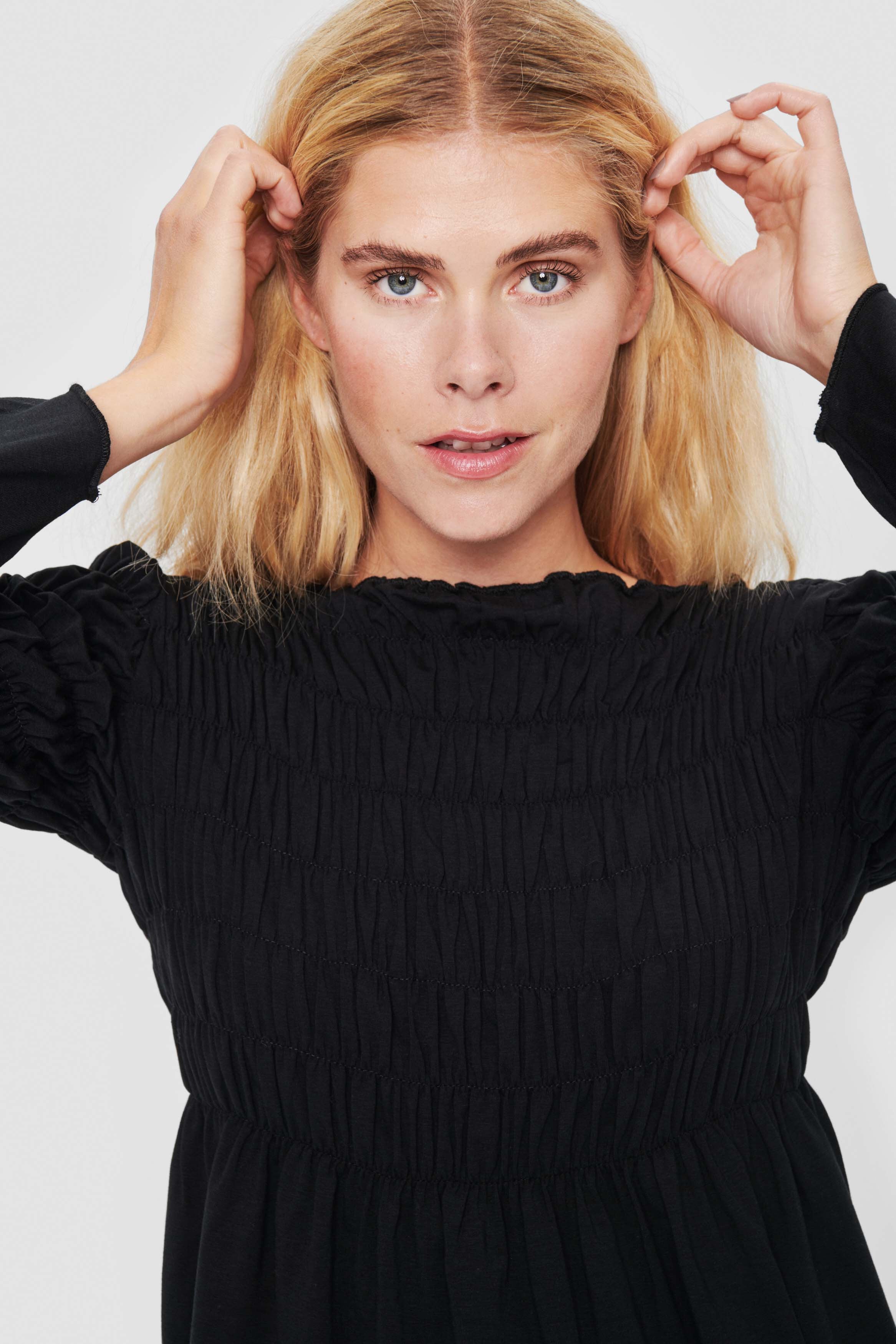 front top half view of a woman wearing the bitte kai rand atlas blouse in black. This blouse features a stitched gathering (smocking) on the top half of the top and sleeves that causes a peplum-like flare out from the waist and elbows.