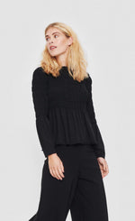 Load image into Gallery viewer, front top half view of a woman wearing the bitte kai rand atlas blouse in black. This blouse features a stitched gathering (smocking) on the top half of the top and sleeves that causes a peplum-like flare out from the waist and elbows.
