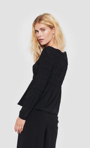 Back top half view of a woman wearing the bitte kai rand atlas blouse in black. This blouse features a stitched gathering (smocking) on the top half of the top and sleeves that causes a peplum-like flare out from the waist and elbows.