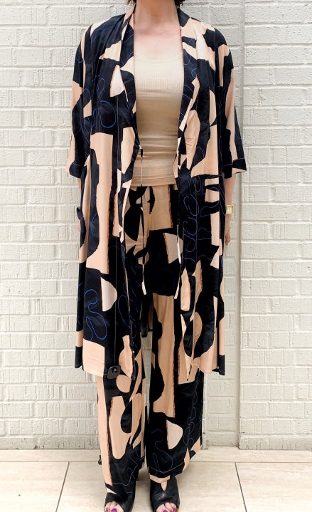 Front full body view of a woman wearing the bitte kai rand monstera wrap dress open over a nude tank and with the bitte kai rand monstera pant. These dress and pants are nude with black and blue abstract print on them. The dress has a tie, 3/4 length sleeves, and ends at the knees.