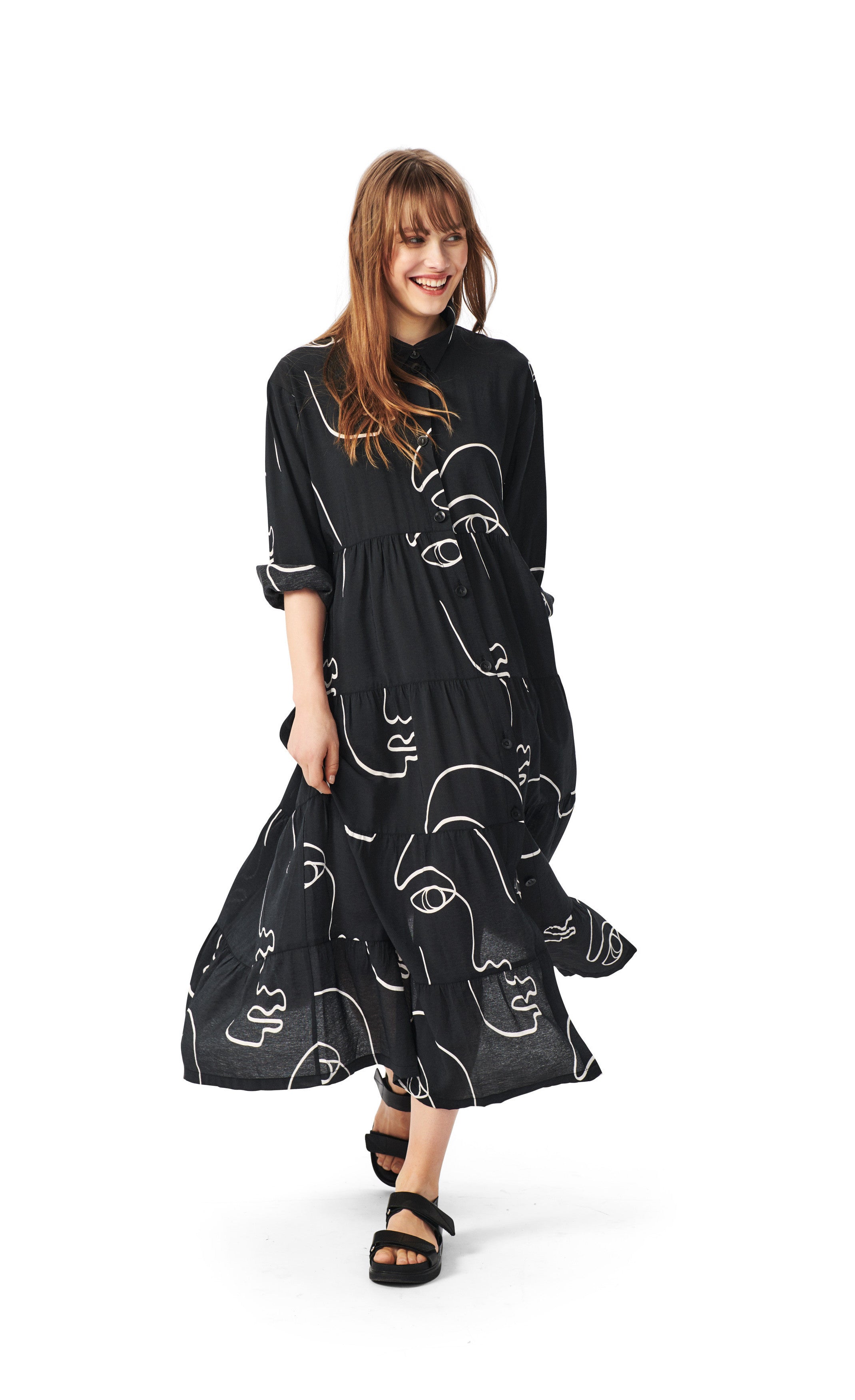 Front full body view of a woman wearing the bitte kai rand sketch viscose long dress. This dress is black with white sketches of faces all over it. The dress has a button up front, a collar, long sleeves that are folded up, and a ruffled bottom half.