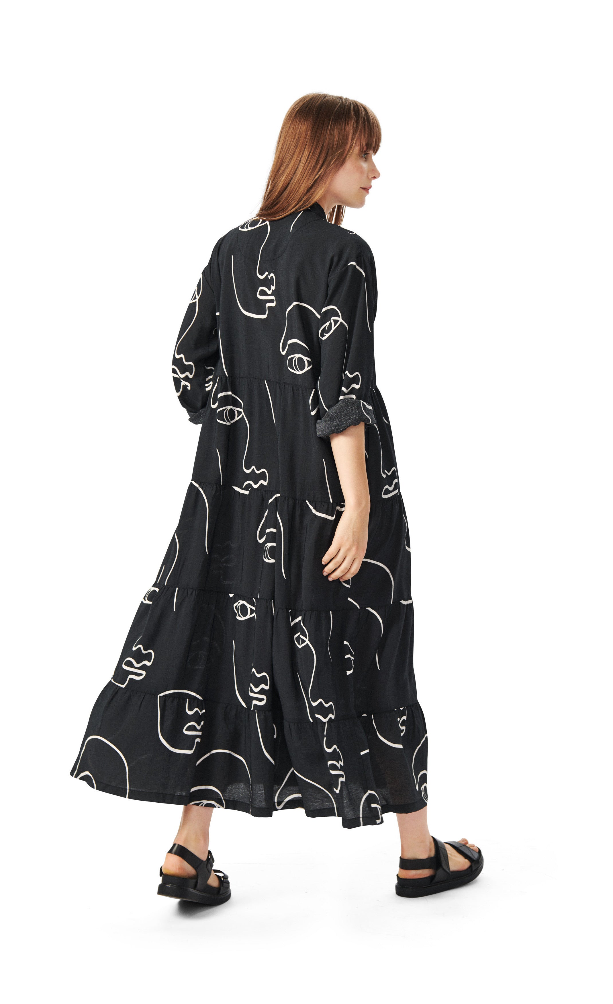 Back full body view of a woman wearing the bitte kai rand sketch viscose long dress. This dress is black with white sketches of faces all over it. The dress has long sleeves that are folded up and a ruffled bottom half.