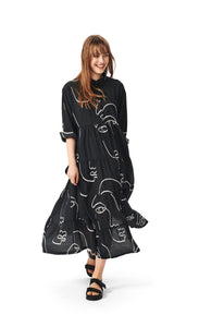 Front full body view of a woman wearing the bitte kai rand sketch viscose long dress. This dress is black with white sketches of faces all over it. The dress has a button up front, a collar, long sleeves that are folded up, and a ruffled bottom half.