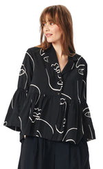 Load image into Gallery viewer, Front top half view of a woman wearing the bitte kai rand sketch viscose shirt jacket. This version of the shirt is black with white sketch faces on it. It has a two button up front, a notched collar, long sleeves with ruffles, and a ruffles on the bottom half of the top.
