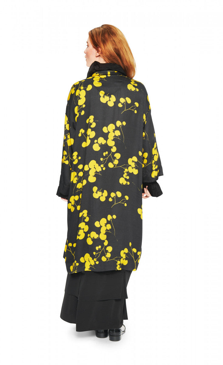Back view of a woman wearing the bitte kai rand winter leaves kimono jacket. This long black jacket has yellow flowers/leaves and an off-center single front button.