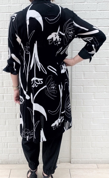 Back full body view of a woman wearing the bitte kai rand tulip tango shirt dress. This dress is black with black and white tulip flowers all over it. The dress has 3/4 length sleeves and ends at the knees.