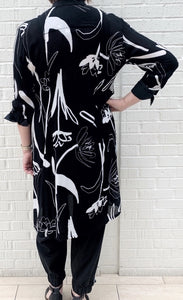 Back full body view of a woman wearing the bitte kai rand tulip tango shirt dress. This dress is black with black and white tulip flowers all over it. The dress has 3/4 length sleeves and ends at the knees.