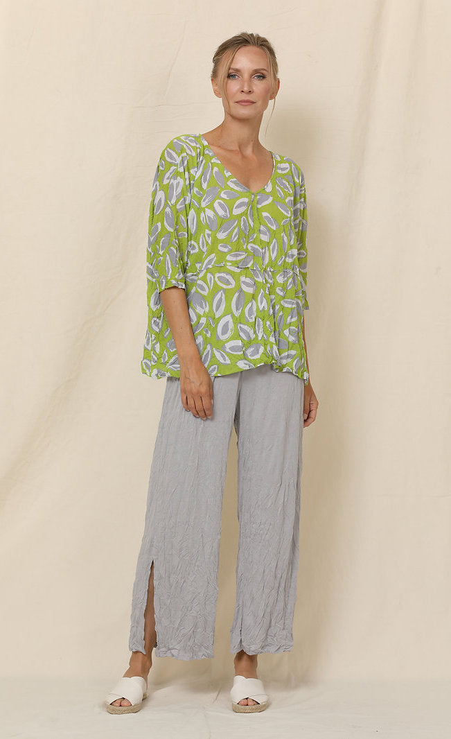 Front full body view of a woman wearing grey crinkled pants and the chalet aimee top. This top is green with grey and white leaves all over it. It has drop shoulder short sleeves, a v-neck, and a boxy fit.