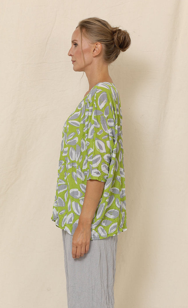 Left side top half view of a woman wearing grey crinkled pants and the chalet aimee top. This top is green with grey and white leaves all over it. It has drop shoulder short sleeves, a v-neck, and a boxy fit.