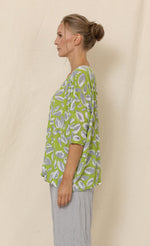 Load image into Gallery viewer, Left side top half view of a woman wearing grey crinkled pants and the chalet aimee top. This top is green with grey and white leaves all over it. It has drop shoulder short sleeves, a v-neck, and a boxy fit.
