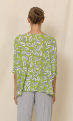 Load image into Gallery viewer, Back top half view of a woman wearing grey crinkled pants and the chalet aimee top. This top is green with grey and white leaves all over it. It has drop shoulder short sleeves, a v-neck, and a boxy fit.
