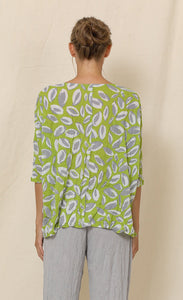 Back top half view of a woman wearing grey crinkled pants and the chalet aimee top. This top is green with grey and white leaves all over it. It has drop shoulder short sleeves, a v-neck, and a boxy fit.