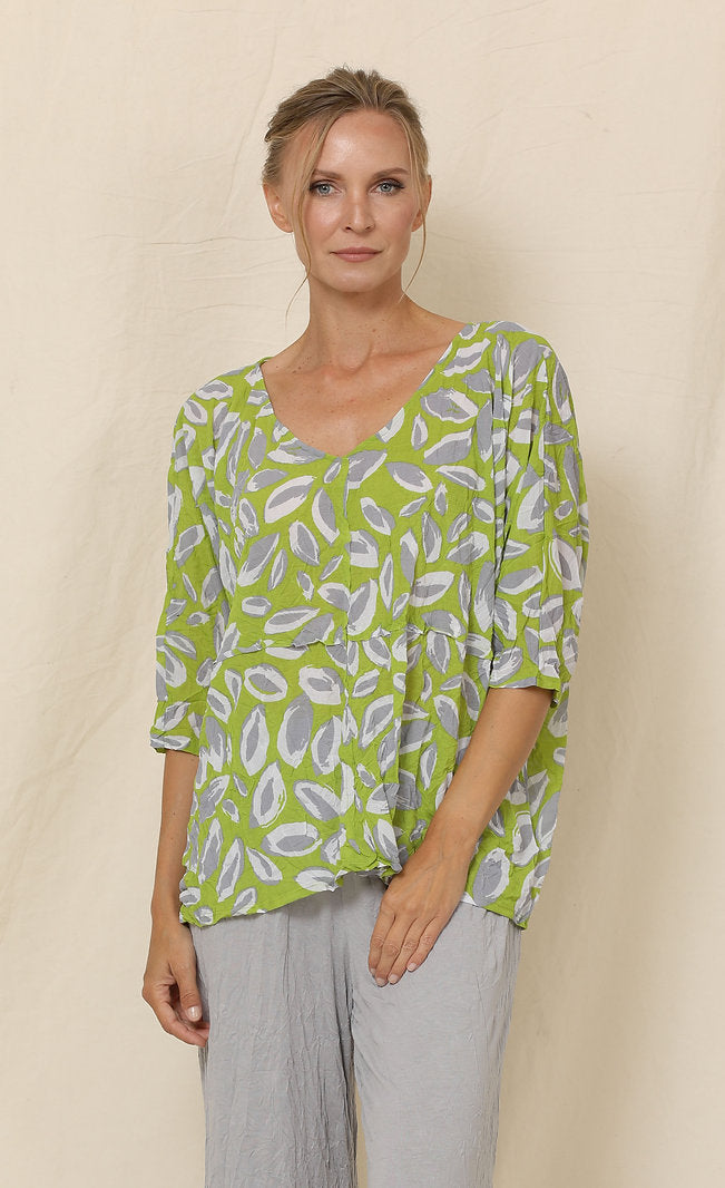 Front top half view of a woman wearing grey crinkled pants and the chalet aimee top. This top is green with grey and white leaves all over it. It has drop shoulder short sleeves, a v-neck, and a boxy fit.