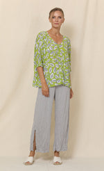 Load image into Gallery viewer, Front full body view of a woman wearing grey crinkled pants and the chalet aimee top. This top is green with grey and white leaves all over it. It has drop shoulder short sleeves, a v-neck, and a boxy fit.
