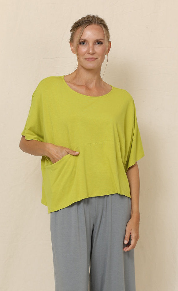 Front top half view of a woman wearing grey pants and the chalet blythe topper in the color moss. This color is a yellow-green. The top has drop shoulder short sleeves, a boxy fit, a single front pocket, and an asymmetrical hem.