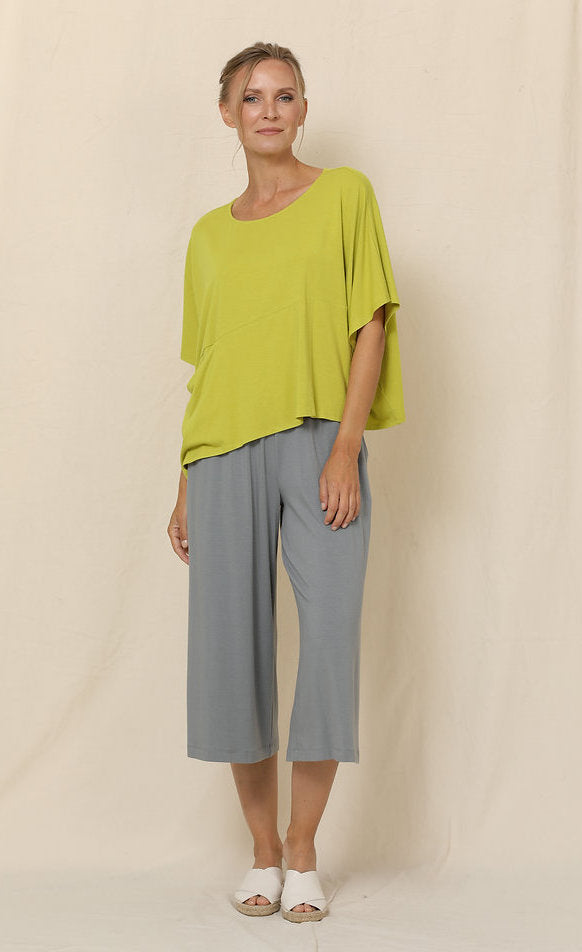 Front full body view of a woman wearing grey pants and the chalet blythe topper in the color moss. This color is a yellow-green. The top has drop shoulder short sleeves, a boxy fit, a single front pocket, and an asymmetrical hem.