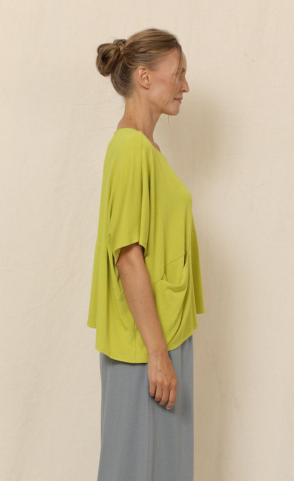 Right side top half view of a woman wearing grey pants and the chalet blythe topper in the color moss. This color is a yellow-green. The top has drop shoulder short sleeves, a boxy fit, a single front pocket, and an asymmetrical hem.