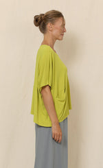 Load image into Gallery viewer, Right side top half view of a woman wearing grey pants and the chalet blythe topper in the color moss. This color is a yellow-green. The top has drop shoulder short sleeves, a boxy fit, a single front pocket, and an asymmetrical hem.
