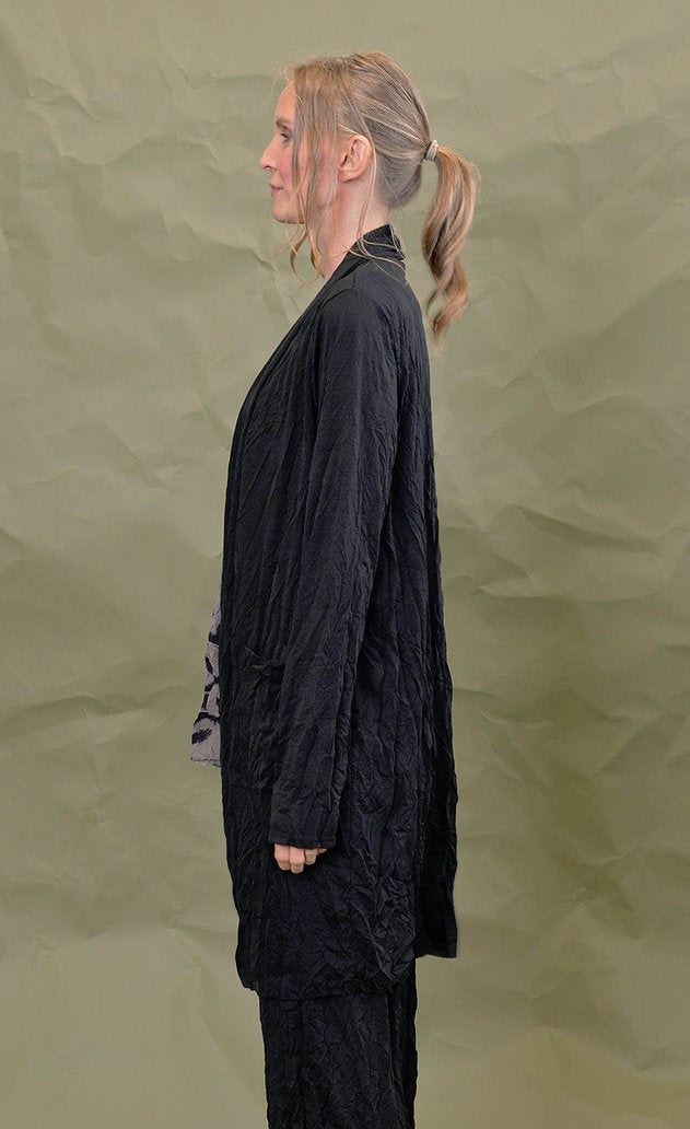 left side top half view of a woman wearing the chalet black cleon cardigan. This long cardigan has long sleeves, a draped open front, and front draped pockets.