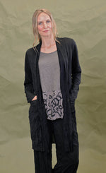 Load image into Gallery viewer, Front top half view of a woman wearing the chalet black cleon cardigan. This long cardigan has long sleeves, a draped open front, and two front draped pockets.
