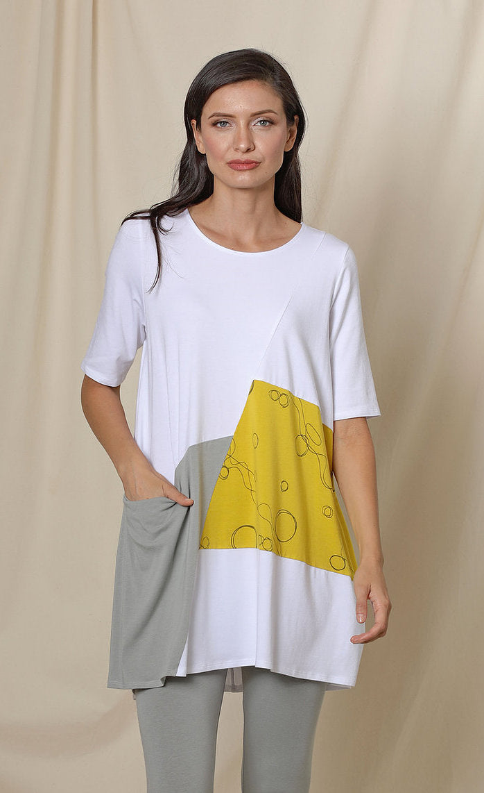 Front top half view of a woman wearing the chalet fallon tunic. This tunic is white with a yellow patch and a grey patch pocket in the front. This tunic sits below the hips.