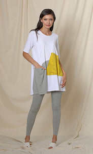 Front full body view of a woman wearing the chalet fallon tunic. This tunic is white with a yellow patch and a grey patch pocket in the front. This tunic sits below the hips.