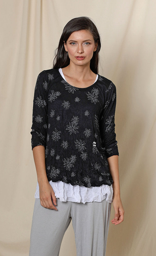 Front top half view of a woman wearing the chalet gizel top. This top is black with white flowers. It has 3/4 length sleeves and a scoop neck.