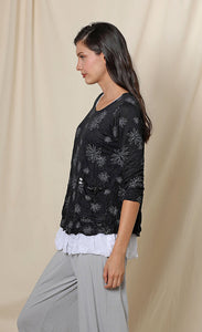 Left side top half view of a woman wearing the chalet gizel top. This top is black with white flowers. It has 3/4 length sleeves, a scoop neck, and a draped pocket on the left side.