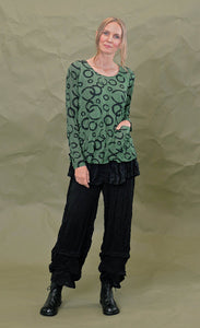 Front full body view of a woman wearing the chalet gizel top in treetop. This top is green colored with black circles. It has long sleeves, a round neck, and a front patch pocket.