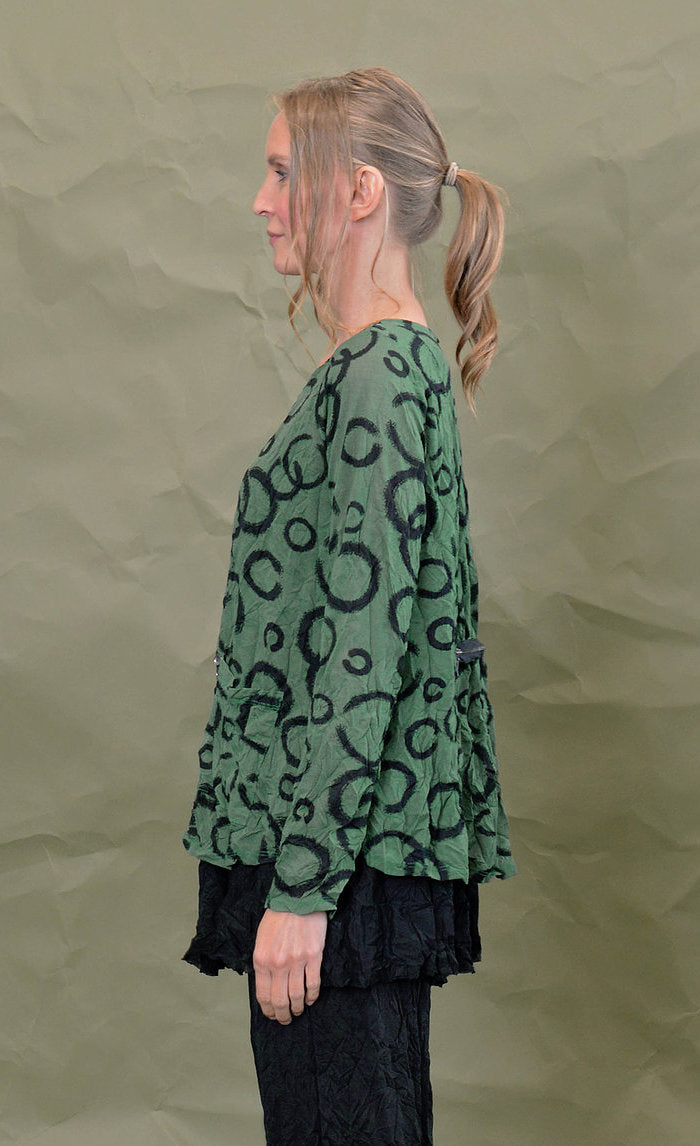 Left side top half view of a woman wearing the chalet gizel top in treetop. This top is green colored with black circles. It has long sleeves, a round neck, and a front patch pocket.
