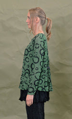 Load image into Gallery viewer, Left side top half view of a woman wearing the chalet gizel top in treetop. This top is green colored with black circles. It has long sleeves, a round neck, and a front patch pocket.
