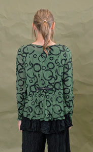 Back top half view of a woman wearing the chalet gizel top in treetop. This top is green colored with black circles. It has long sleeves and a decorative zipper on the back that gathers the fabric.