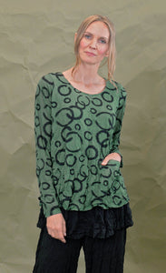 Front top half view of a woman wearing the chalet gizel top in treetop. This top is green colored with black circles. It has long sleeves, a round neck, and a front patch pocket.