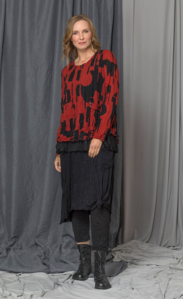 front full body view of the chalet gizel top. This top is red with a black retro print. It has long sleeves, a round neck, a front draped pocket, and crinkled fabric.