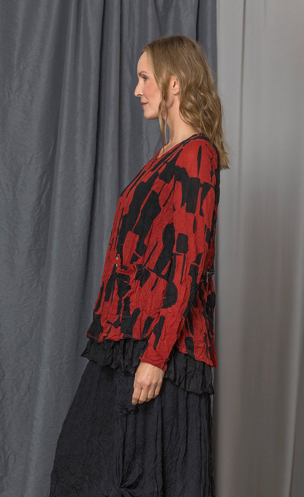 left side top half view of the chalet gizel top. This top is red with a black retro print. It has long sleeves, a round neck, a front draped pocket, and crinkled fabric.
