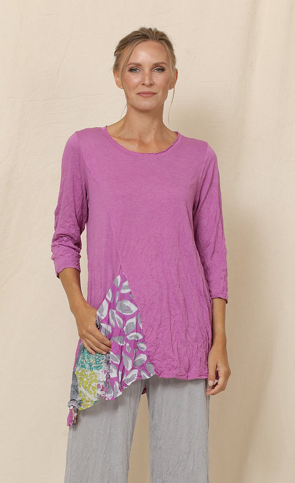 Front top half view of a woman wearing grey crinkled pants and the chalet mulberry greta tunic. This tunic is pink with 3/4 length sleeves, a scoop neck, and an asymmetrical pointed hem. The front of the top has a draped printed pocket on the right side.
