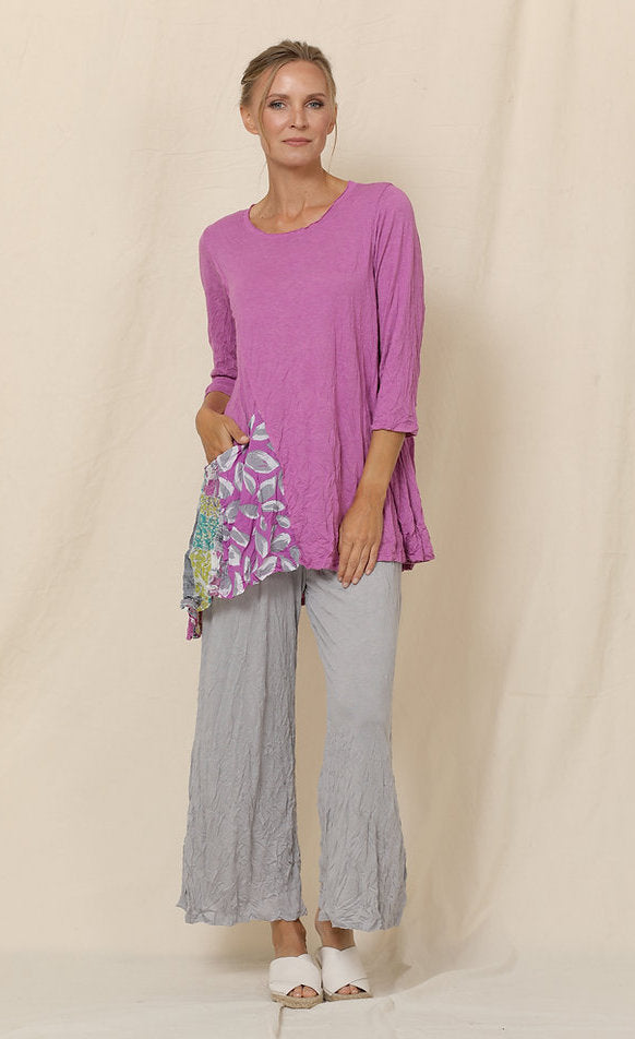 Front full body view of a woman wearing grey crinkled pants and the chalet mulberry greta tunic. This tunic is pink with 3/4 length sleeves, a scoop neck, and an asymmetrical pointed hem. The front of the top has a draped printed pocket on the right side.