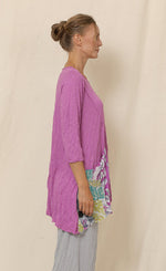Load image into Gallery viewer, Right side top half view of a woman wearing grey crinkled pants and the chalet mulberry greta tunic. This tunic is pink with 3/4 length sleeves, a scoop neck, and an asymmetrical pointed hem. The front of the top has a draped printed pocket on the right side.
