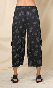Back bottom half view of a woman wearing the chalet hallie pant. This pant is black with white flowers. It has a crinkled look and two draped side pockets. The pants end above the ankles.