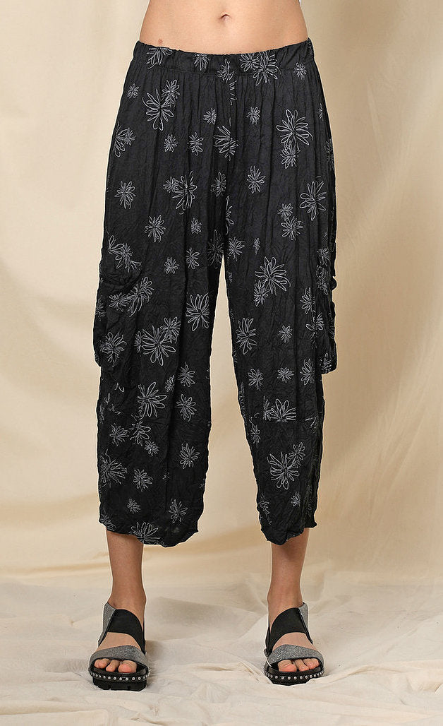 Front bottom half view of a woman wearing the chalet hallie pant. This pant is black with white flowers. It has a crinkled look and two draped side pockets. The pants end above the ankles.