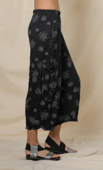 Load image into Gallery viewer, Right side, bottom half view of a woman wearing the chalet hallie pant. This pant is black with white flowers. It has a crinkled look and two draped side pockets. The pants end above the ankles.

