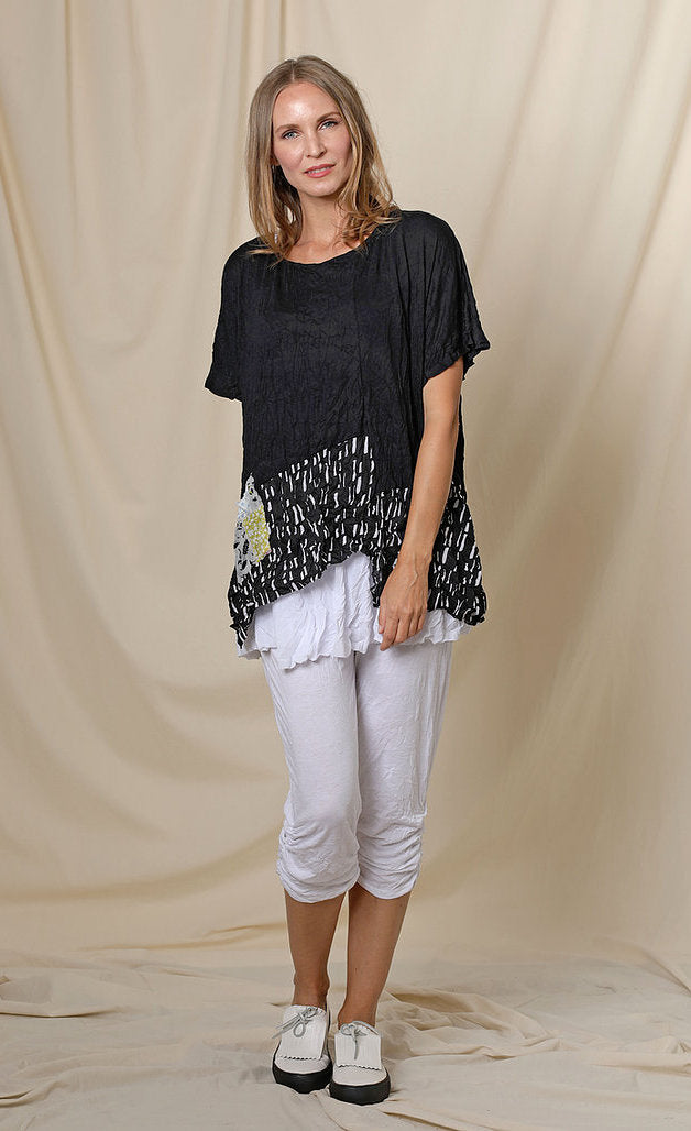 Front full body view of a woman wearing the chalet josephine top. This short sleeve top is black with a black and white print band at the bottom and a patchwork printed front pocket.